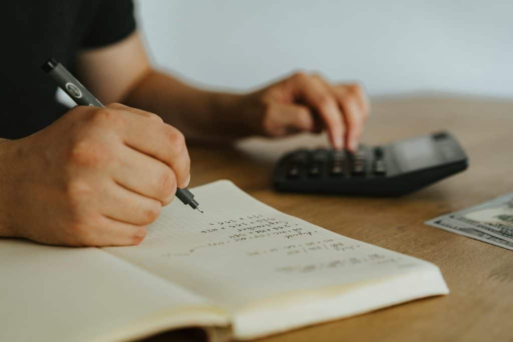 Photo of somebody sitting at a desk. You can only see their hands and they're doing their taxes. They have a notebook and a calculator out.